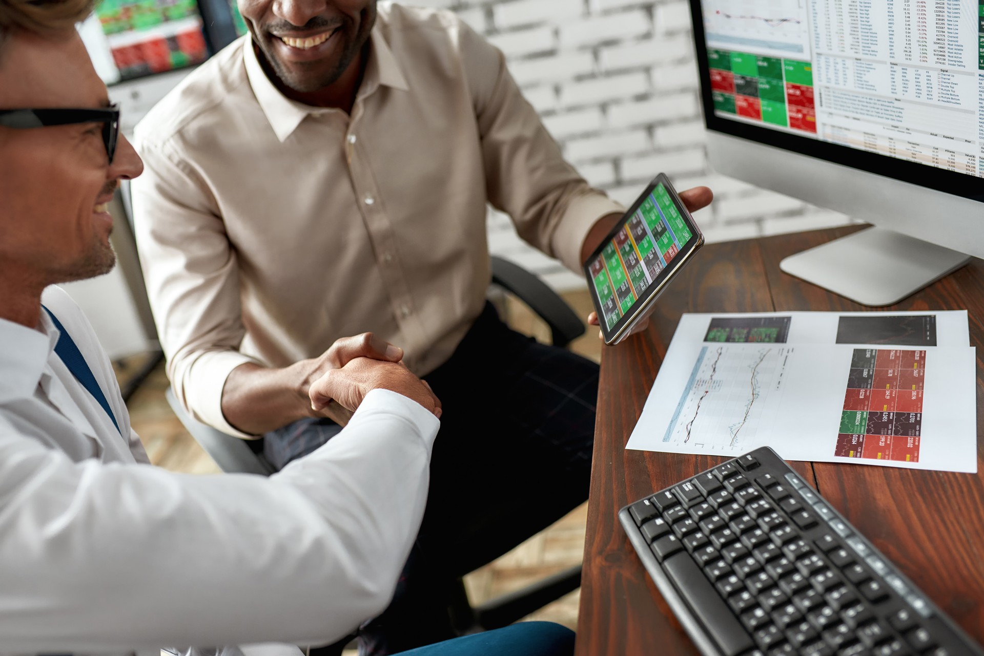 We will do it. Business handshake. Two businessmen, traders reaching an agreement and making a deal. Monitor with charts, data analyses in background